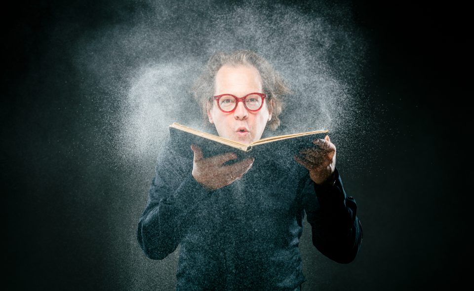 Norbert Trawöger entstaubt mit einem Lufthauch ein aufgeschlagenes Buch, das er in beiden Händen hält mit Blick in die Kamera.